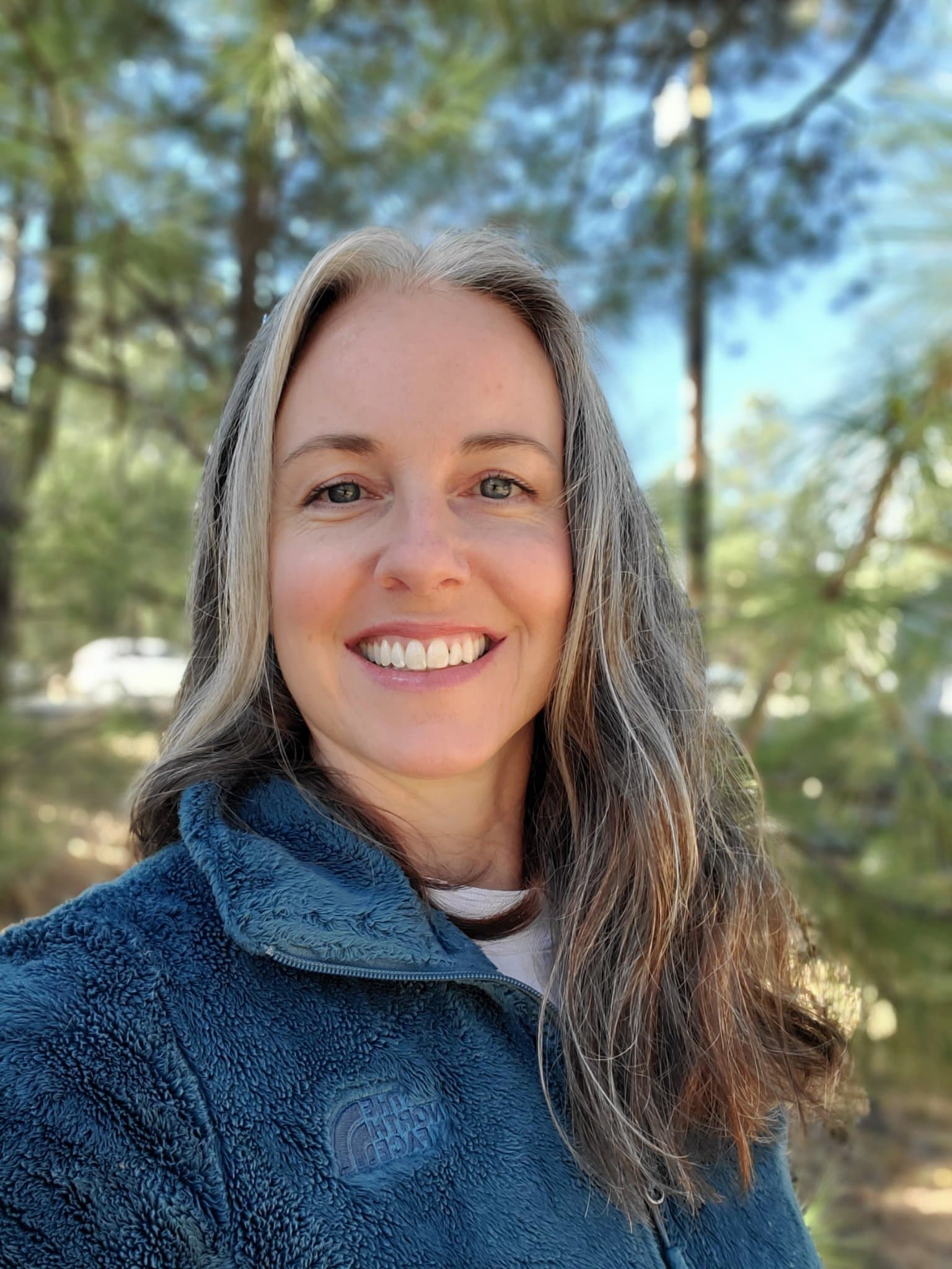 Portrait of Melony Peters, Content Manager of Stroll St. Pete, smiling outdoors in a teal fleece jacket among pine trees